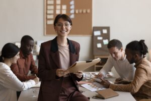 Gestora sorrindo com prancheta, equipe trabalhando ao fundo, ilustrando os pilares da liderança eficaz e alta performance com Agregarh.