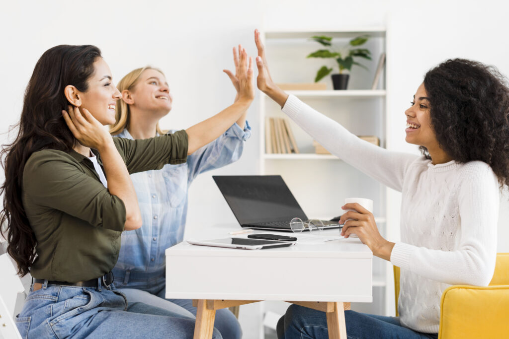 A imagem mostra três mulheres sentadas em volta de uma mesa, tocando suas mãos e sorrindo. O ambiente é corporativo.